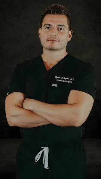 Male healthcare professional in black scrubs with arms crossed, posed against a dark background for a portrait.